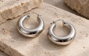 Editorial illustration: Two large hollow-style stainless steel hoop earrings on a neutral stone surface, complete realistic products